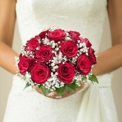 A radiant snapshot of bridal anticipation, this floral portrait centres on a bride's hands-her fingers delicately encircling a domed bouquet that sings with the deep passion of red roses. Each rose appears lush and tightly woven, their velvet texture and subtle gradations of crimson symbolising enduring love. Tucked between the roses, baby's breath unfurls in airy, white constellations-softening the robust palette and offering the lightness often seen in bouquets leaving the doors of North Watford's local flower shops. At the margins of the bouquet, a few fresh green leaves emerge, their hue reminiscent of Oxhey Park's springtime, infusing the arrangement with vitality. The bride's ivory gown, glimpsed behind the blooms, features an intricate lace or embroidered detail across the bodice, evoking the elegance of a Watford wedding venue. Flows of pale fabric trail along her side, their movement echoing the gentle rhythm of a riverside breeze. In the background, the cream and beige tones blur to silence, allowing the bouquet's artistry to speak for itself. Here, tradition meets artistry; the floral design feels both classic and deeply personal-as if paired to celebrate every joyful promise made close to North Watford High Street. This bouquet, steeped in fragrance and romance, promises to be remembered long after the last petal has fallen.