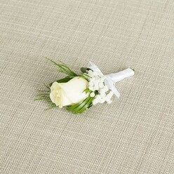 A refined wedding boutonniere is displayed at a gentle angle on a neutral, light beige background that looks like natural linen, its fine crosshatch texture adding an understated, almost rustic elegance reminiscent of intimate receptions in North Watford's characterful halls and restaurants. At the centre rests a single creamy white rosebud, its petals smooth and velvety, folded in tight spirals that suggest both purity and the promise of a day just beginning. Around this focal bloom, clusters of soft white baby's breath create a misty, cloud-like frame, each tiny blossom introducing a sense of airiness and movement, as if a light breeze were passing through. Slender, vibrant green leaves and a few feathery, fern-like sprigs curve naturally along the sides of the rose, introducing varied shades of green that echo the foliage of nearby parks and gardens, offering a fresh, woodland note to the composition. The stems are meticulously bound in a band of white satin ribbon, its surface smooth and faintly luminous, with a small, symmetrical bow tied just below the floral head, indicating the care and precision of a skilled florist accustomed to dressing grooms and groomsmen for ceremonies around North Watford. Soft, even lighting falls across the scene, revealing the delicate texture of the petals, the tiny globes of gypsophila flowers, and the subtle sheen of the ribbon without any glare, giving the piece a calm, contemplative aura. This boutonniere feels perfectly suited to a groom stepping out from a terraced home off St Albans Road or arriving at a local venue, its quiet elegance mirroring the gentle excitement and heartfelt commitment of a North Watford wedding day.