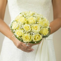 Beneath the gentle glow of a North Watford afternoon, this striking bridal bouquet is held gracefully against the elegant white expanse of a wedding gown, thought to be crafted in the style beloved by local brides. Each soft yellow rose, tenderly selected for its fullness and flawless spirals, is gathered with devotion, creating a rounded cluster that appears both lush and tranquil. Scattered throughout the bouquet, sprigs of snowy baby's breath-known to florists in Watford Market as a symbol of purity and hope-add a cloud-like quality, breaking up the golden hues with their airy, delicate charm. At the bouquet's base, subtle rings of lustrous greenery or artfully entwined wire gather the stems in a neat, contemporary finish, their deep tones highlighting the bouquet's graceful form. The hands cradling this arrangement glimmer with barely-there polish and the sparkle of a wedding ring, reinforcing the tenderness and anticipation unique to wedding mornings near Cassiobury Park. The background, a wash of gentle neutrals and softened textures, allows the bouquet to take centre stage-exuding freshness, care, and the artistry associated with Flower Delivery North Watford. Perfect for those seeking a romantic and classic floral centrepiece for their big day, this image perfectly captures the scent of blooming roses and the essence of North Watford's warm, community-driven celebrations.