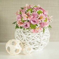 Imagine stepping into a bright Watford home near Cassiobury Park and discovering this stunning floral centerpiece waiting to greet you with a sense of poised tranquility. A smooth, white open-weave vase, its design echoing the handiwork of local artisans, anchors an abundant bouquet overflowing with pastel pink and lavender roses, the plushness of their petals softening the mood. Pink alstroemeria, with delicate striped blooms, suggest quiet congratulations and thoughtful gatherings-perhaps for an intimate North Watford anniversary or birthday. Lime green button chrysanthemums, round and cheerful, punctuate the flowerscape with texture, complemented by sprigs of pristine white baby's breath that lend an airy, floating quality reminiscent of spring clouds above the Grand Union Canal. Deep, rich green leaves peek through the arrangement, providing a visual grounding much like the leafy borders of Watford's neighbourhoods. To one side, two decorative natural-toned spheres-evoking carefully woven rattan crafts-bring balance and a contemporary accent reminiscent of Watford's blend of tradition and modernity. The background, a soft beige with subtle texture, frames the composition gently, allowing the bouquet's colors and forms to glow with composed energy. Perfect as a wedding table piece or an elegant gift, this centerpiece tells a story of thoughtful design, heartfelt celebration, and North Watford's signature blend of warmth and creative expression.