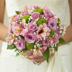 In the heart of North Watford, a bride's bouquet is brought to life by artisan hands, uniting tradition and modern flourish. Cradled delicately against an out-of-focus lace wedding dress, the bouquet's visual poetry is immediately captivating-roses in gentle shades of pink and lavender reflect the soft pastels of spring mornings by Watford Fields, their blooms at once timeless and touched with whimsy. The crisp lime button chrysanthemums, perfectly spherical and bursting with life, offer a fresh vibrance that dances alongside the smooth rose petals. Each pink alstroemeria dazzles with streaks of magenta and sunlit yellow, contributing an air of playful sophistication and subtle scent that lifts the bouquet's spirit. Artful sprays of white baby's breath settle like mist, blurring the boundaries between each flower and lending a sense of lightness akin to a dawn fog rolling over Oxhey Park. Dark green foliage gives structure and depth, tying together a look that feels both classic and distinctly contemporary. To hold such a bouquet in North Watford is to carry a piece of the local landscape and a story of joyous commitment, a marriage of colour and form thoughtfully tailored for heartfelt celebrations and elegant affairs alike.