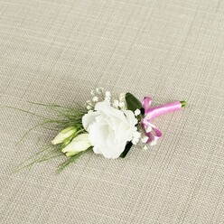 An elegant floral boutonnière is photographed from a slightly raised angle, resting diagonally on a warm, light beige textile that resembles hand-loomed linen, the weave clearly visible and adding rustic charm. In the centre of the composition, a fully opened white lisianthus bloom displays its intricate, ruffled petals, each layer curling softly outward to create a rounded, almost rose-like form that feels serene and polished. Nestled just beneath and to the left of this focal flower are two unopened lisianthus buds in a tender, creamy green, their slender shapes suggesting the quiet anticipation that often surrounds a wedding morning in North Watford. Scattered delicately through the arrangement are airy tufts of tiny white gypsophila, their minute blossoms forming a sparkling halo around the main flower and lending the buttonhole an ethereal, cloud-soft texture. From behind the lisianthus, fine strands of dark green, grass-like foliage extend gracefully towards the bottom left, adding movement and an organic, garden-picked feel, while a single, broader dark green leaf sits behind the bloom, anchoring the design with deeper colour. At the lower right, the stems are crisp and neatly aligned, bound together by a vivid fuchsia satin ribbon tied into a jaunty, compact bow, its glossy surface catching the light and offering a cheerful burst of colour against the neutral background. The combination of white, green, and bright pink suggests a joyful, contemporary celebration, perfect for a groom, usher, or guest heading to a ceremony near St Albans Road or the quieter residential streets of North Watford. The overall impression is one of fresh refinement, subtle fragrance, and handcrafted care, the kind of finely detailed piece a local florist would prepare just before same-day delivery.