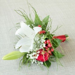This exquisite North Watford bouquet, elegantly presented on a softly textured, creamy linen surface, features a central white lily whose unfurled petals showcase faint russet speckles and golden anthers, radiating purity and tranquil allure. The arrangement is complemented by a deeply coloured red rose, its opulent bloom representing love and devotion. Tracing through the composition, clusters of bright red berries gleam like joyful notes, reminiscent of festive market stalls in North Watford during the holiday season. Sprigs of fine baby's breath add gentle clouds of white, softening the display and capturing the feel of wildflowers found along Watford's leafy avenues. Interlaced throughout are delicate green fern fronds and broad, glossy leaves, grounding the piece with natural abundance and texture. At the edge, a poised, light green lily bud stretches forward, signalling growth and new beginnings. The bouquet's stems are gathered with a silky red satin ribbon, artfully knotted to bring a touch of refined celebration and echo the crimson highlights. Meticulously composed by North Watford's skilled florists, this compact floral design bridges classic romance with contemporary festivity, making it a perfect choice for bridesmaids, local weddings, milestone events, or as a heartfelt gesture for someone special in the community-all while reflecting the artisanal spirit of Flower Delivery North Watford.