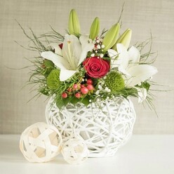 A sophisticated floral centrepiece sits poised on a clean white tabletop, the kind you might find in a beautifully dressed wedding venue around North Watford. At its heart is a distinctive round vase in matte white, crafted from interlaced strands that mimic woven wicker or wrapped yarn, giving it a light, sculptural quality. The vase cradles a full arrangement of fresh flowers in a timeless white, red, and green palette. Large white lilies dominate the composition, their trumpet-shaped blooms opening wide to display fine speckling and deep yellow stamens, while several unopened buds rise gracefully above, hinting at the arrangement's continued life over the coming days. Nestled closely to the lilies are deep red roses, each petal velvety and gently curved, with one central rose drawing the eye as a focal point of passion and romance. Rounded tufts of bright green pompon blooms, probably green dianthus or chrysanthemums, provide playful spherical accents and a vivid splash of colour between the white petals and darker foliage. Fine sprays of baby's breath thread through the bouquet like tiny stars, softening the bold hues, while scattered red berries add an almost festive sparkle, perfect for winter celebrations or a Christmas-time wedding breakfast near Garston or along St Albans Road. Slender fronds of asparagus fern and other mixed greenery spill outward, giving the design a slightly untamed, natural character without losing its elegance. Beside the vase, two decorative woven balls in a pale timber shade echo the texture of the container and complete the styled scene. The neutral, softly textured background allows every delicate detail of the flowers to stand out, creating a serene yet celebratory mood ideal for wedding tables, engagement dinners, or special family gatherings in North Watford.