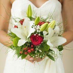 The artistry of a North Watford wedding comes to life in this close-up of a bridal bouquet, cradled lovingly by the bride in her gracefully textured white gown. Luminous white oriental lilies open wide among the blooms, their golden stamens catching the ambient light that fills the room near Watford's tranquil parks. Between these lilies, deep red roses spiral in passionate contrast, their velvety richness drawing the eye and evoking lifelong commitment. Vivid red hypericum berries, bright and cheerful, bring playful festive notes that recall local celebrations along Courtlands Drive. Sprigs of delicate white baby's breath lend a whimsical, airy texture, floating like morning mist through the bouquet. Graceful, unopened green lily buds are nestled thoughtfully, promising beauty and happiness in days yet to come. Mossy green button flowers, slightly spherical and unique, add depth and a touch of the wild North Watford greenery, while a variety of ferns and smooth leaves complete the lush base. The composition feels both classic and contemporary, echoing the diverse spirit of the community. Beyond a mere bouquet, this creation is alive with meaning-a tactile expression of hope, love, and artistry, woven together by experienced local florists. Imbued with gentle fragrance, subtle textures, and memories in the making, it's a tribute to joy and the traditions cherished in North Watford weddings.