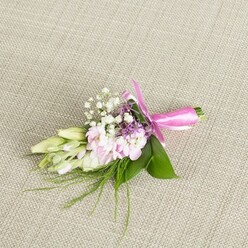 A finely detailed boutonniere or petite corsage lies gently on a pale beige, linen-like fabric, its woven texture softly visible beneath the stems and ribbon, evoking the relaxed elegance of a North Watford gathering. At the heart of the design sits a close cluster of blush and soft pink blossoms, their many ruffled petals layered in delicate folds that give a sense of movement, as if they've just been picked from a garden on a quiet side street off St Albans Road. Tiny sprays of classic white baby's breath weave between the pink flowers, their airy shapes creating a lace-like veil that softens every edge. Tucked discreetly among them are a few small lavender and deep purple florets, adding depth and a cool, evening-sky tone without disturbing the gentle palette. To one side, a line of slender buds in creamy white and pale green unfurls along the length of the arrangement, suggesting freesia or similar buds about to bloom, and hinting at the promise of the celebrations ahead. Broad, glossy green leaves cradle the central cluster, while fine, feathery strands of grass-like foliage extend outward, bringing a fresh, garden-grown feeling typical of local florals arranged near Garston or Park Avenue. The stems are wrapped neatly in a vivid fuchsia satin ribbon, tied into a symmetrical bow whose smooth loops and slightly trailing ends catch the light. Every element-from the subtle sheen of the ribbon to the cool, velvety petals-communicates thoughtful craftsmanship, making this piece an ideal finishing touch for a wedding, prom, or graduation photos taken around North Watford, where understated style and heartfelt detail matter most.