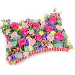 Seen from a slightly raised, angled viewpoint on a pristine white plane, this rectangular floral pillow is an intensely crafted statement of remembrance, assembled with a florist's attention to proportion and feel. The top plane is a compact meadow of hot pink roses-some in tight bud, others opening to reveal layered petals-interlaced with pale pink, frilly carnations that give a soft, tactile counterpoint. Lemon-yellow ranunculus provide bright, cheerful beads of colour, while purple lisianthus add cooler, bell-like tones that deepen the visual field. Blue eryngium thistles reintroduce a silvered, spiky texture, their sculptural silhouettes breaking the otherwise rounded language of the arrangement. Eucalyptus leaves, round and silvery-green, and additional lush foliage are worked throughout to create depth, gentle scent, and a natural underpinning that allows each bloom to read clearly. The perimeter is finished with a deep red pleated or ruffled satin ribbon that forms a defined edge-an elegant frame for the cushion's floral carpet and a nod to traditional memorial styling. The composition was designed to comfort: the lively yet soft palette suggests celebration of life, and the density of blooms offers a visual embrace. As a local piece from Flower Delivery North Watford, it's the kind of memorial cushion we prepare for services near Cassiobury Park and on Watford High Street, chosen by families who want a tribute that expresses warmth, enduring affection, and careful craftsmanship.