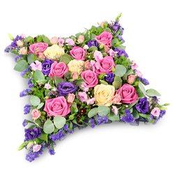 Set against a bright white backdrop, a luxurious floral cushion in the shape of a soft-edged rectangle seems to float, its rounded corners and plush density offering a visual sense of rest and release. The tribute is composed of tightly nestled roses, lisianthus, and filler flowers in a palette of warm pinks, creamy yellows, and rich purples, reminiscent of a serene garden on a still afternoon in North Watford. Large, open roses in vivid fuchsia and gentle pastel pink form the heart of the arrangement, their velvety petals spiralling outwards in perfect rosettes. Interspersed among them are pale yellow and cream-toned roses, their more delicate hues lending calm balance to the stronger pinks. Deep purple lisianthus blooms punctuate the design, their ruffled, slightly folded petals adding a velvety texture and a sense of depth, like shaded corners under mature trees along nearby residential streets. Between these hero flowers, clusters of tiny lavender and lilac blossoms, likely statice or limonium, appear as fine, cloudlike sprigs filling every gap with soft colour. Encasing the flowers is a cushion of mixed greenery: rounded, blue-green eucalyptus leaves bring a cool, silvery note, while more slender, ferny foliage offers structure and a darker green counterpoint. Every stem seems deliberately placed, reflecting the practiced hand of a local funeral florist familiar with creating tributes that speak where words fail. This floral pillow would be a fitting sympathy piece for a chapel, crematorium, or church service in North Watford, expressing love, gratitude, and gentle farewell.