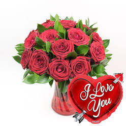 An opulent display of romance dominates this image: a clear, ruby-red fluted glass vase brimming with lush, deep red roses and fresh green foliage, styled with the care of an experienced North Watford florist. The roses are densely packed, creating a rich dome of colour, with some blooms fully open and revealing their layered, velvety petals, while others remain partially closed, hinting at days of beauty still to come. The petals have that soft, almost satin texture that invites you to imagine their gentle fragrance filling a cosy home near St Albans Road or along the quieter residential streets of North Watford. Interwoven between the roses, dark green leaves and stems provide structure and contrast, their slightly glossy surfaces catching the light. The vase itself has a graceful, fluted shape, its translucent red glass adding depth as it holds the stems in perfectly balanced proportions. In the lower right corner, angled towards the viewer, a bright red heart-shaped foil balloon leans into the scene. It is highly reflective, with highlights dancing across its surface, and it bears the bold white cursive words I Love You, framed by a silvery arrow shooting diagonally through the heart. Set against a pure white background, every shade of red appears more intense, every green leaf more vivid. The arrangement feels like a romantic gesture ready for same-day delivery across North Watford, ideal for anniversaries, proposals, or a spontaneous declaration of love that feels both heartfelt and memorable.