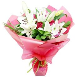 A luxurious bouquet of pristine white lilies and rich red roses stands out dramatically against a clear white background, as if placed on a light-filled counter in a North Watford flower shop. The composition is dominated by several open Oriental lily blooms, their star-like, trumpet petals unfolding in pure, creamy white and curving back to reveal prominent, dark chocolate-brown stamens dusted with pollen. Close by, some lilies remain in bud, sheathed in soft green with hints of ivory at the tips, suggesting that the bouquet will continue to change and open over the coming days. Threaded between the lilies, clusters of deep red roses provide a passionate contrast in both colour and texture; many are still tightly furled, their velvety petals overlapping in perfect spirals that speak of classic romance. Encircling these focal blooms is an abundance of dark green foliage, from broad, lacquered leaves to finer, blade-like greens that arc outward, adding height and natural movement reminiscent of the greenery lining North Watford's residential gardens. The florist has wrapped the stems in a wide layer of bright pink, glossy cellophane, which catches and reflects the light in undulating folds, creating a playful yet refined frame around the flowers. At the base, a slim lime green ribbon is tied neatly in a small bow, its fresh citrus tone complementing the foliage and gently lifting the entire colour palette. The finished bouquet feels celebratory and heartfelt, a sophisticated choice for same-day delivery across North Watford to celebrate anniversaries, special milestones, or to send a warm, affectionate message to someone along the routes toward Garston or Leavesden.