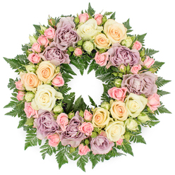 Centered against a clean, white backdrop, a luxurious circular funeral wreath appears almost like a soft pastel halo, its flowers packed closely together to form a continuous tapestry of petals. This memorial design is dominated by roses in varying stages of bloom: large, open-faced ivory and creamy white roses displaying layers of velvety petals; clusters of blush pink spray roses tucked between them, some still in bud, others half-open with shy, curled edges. Hints of pale peach and buttery yellow weave warmth through the composition, while distinctive dusty lavender and muted mauve roses introduce a subtle, vintage note that deepens the overall colour story. The base is built from lush, dark green fern fronds and similar finely cut foliage that peek out around the roses, adding texture and contrast without overwhelming the softness of the blooms. The wreath has a harmonious, well-balanced feel, like something you might see resting gently beside a memorial in North Watford or at a quiet service near Garston and Leavesden. Its round, unbroken form suggests the cycle of life and the enduring nature of love, while the gentle colours evoke peace, comfort, and quiet reflection. Crafted by local florists familiar with the sensitivities of farewell services in the North Watford community, this floral tribute feels both personal and respectful, a serene expression of sympathy and support for families saying goodbye.