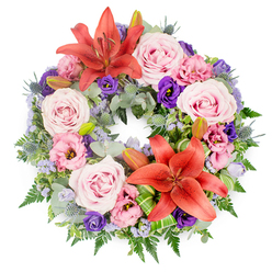 Centred against a pure white background, this sympathy wreath appears like a carefully painted circle of flowers, each bloom chosen to express tenderness and quiet strength. At first glance, the eye is drawn to two bold coral-orange Asiatic lilies placed opposite each other along the ring, their open petals and dusted stamens symbolising courage and heartfelt devotion. Surrounding them is a generous cluster of pastel roses in soft blush and lavender-pink, their gently spiralling petals creating a sense of calm and intimacy. Nestled between the roses are lisianthus in both delicate pink and deep purple tones, their ruffled, cup-shaped blossoms adding volume and a touch of gentle drama. Fine sprays of lavender-blue filler flowers, similar to statice or limonium, blur the transitions between larger blooms, like a light mist of colour. The greenery beneath is abundant and varied: silvery-green eucalyptus leaves, feathery dark green fern fronds, and distinctive spiky blue eryngium thistles, all working together to frame the floral circle with texture and depth. Some bright green, artistically folded leaves are placed near the lower lily, suggesting the hand of an experienced florist. A few unopened lily buds sit thoughtfully among the foliage, evocative of memories still cherished. This wreath would be at home in a quiet chapel or crematorium around North Watford, offering a gentle, dignified expression of sympathy for families seeking a floral tribute that feels both personal and refined.