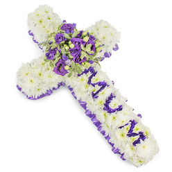 This cross-shaped sympathy arrangement sits alone on a neutral white field, its structure formed from countless small white chrysanthemums, each bloom placed tightly to create a plush, cloud-like surface that reads as purity and repose. A ruffled band of purple satin ribbon-pleated and slightly lustrous-outlines the whole cross, providing a composed frame that draws the eye inward. Down the centre of the vertical bar, the word MUM is crafted in vivid purple, letters composed of compact purple blooms such as lisianthus or petite spray chrysanthemums; the name declares personal loss with tenderness and clarity against the white background. Anchoring the design at the arm intersection is a concentrated focal spray: deep violet roses with curled centers, pale lavender lisianthus, and soft eustoma mingle with variegated green foliage-leaves edged in pale lime with darker centres-and a few glossy green berries that add a gentle, tactile contrast. The combined textures-silken ribbon, velvety roses, powdery chrysanthemums and crisp variegated leaves-make the piece feel thoughtfully weighted and consoling. Under soft diffused lighting the purples read from pale mauve to near-plum, evoking dignity and calm rather than theatricality. The arrangement carries a light floral perfume, subtle and clean, suited to a chapel or funeral parlour. As an experienced florist serving North Watford neighbourhoods from Watford Junction to the avenues near Vicarage Road, Flower Delivery North Watford prepares this kind of tribute with sensitivity for wakes, memorial services, and graveside farewells, offering careful hand-delivery across the area.