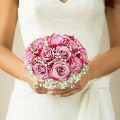 This photograph reveals a bride from North Watford holding a bouquet that radiates joy and timeless beauty, nestled gently against her white, lace-embellished wedding dress. The roses are the heart of the arrangement, each bloom showcasing a seamless gradient from a delicate blush pink at the center to a bold magenta on the outer petals. Such striking colour transition lends the bouquet remarkable depth, drawing the eye to the craftsmanship expected from a seasoned North Watford florist. Dotted generously among the roses, frothy clusters of white baby's breath bestow a soft, almost dewy halo, echoing the fresh beginnings of a wedding day morning. The bride's hands, nuanced with soft, natural manicures, embrace the bouquet just below a sash detail, their warmth and poise accentuating the emotional weight of the day. Subtle hints of green foliage peek from the tight, neatly gathered stems - a quiet nod to gardens along Nascot Wood or leafy Watford Fields. The softly blurred, neutral background allows the vibrancy of the flowers and the lacework of the dress to take centre stage, inviting thoughts of local celebrations and cherished memories made in North Watford. This floral creation is more than a bouquet; it's a tangible chapter in a story of love, artistry, and the gentle hopes woven into every blossom.