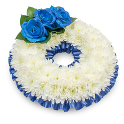 Captured against a pristine white background from a slight overhead angle, this traditional circular wreath is composed of hundreds of pure white chrysanthemums tightly arranged to form a soft, textured ring. The chrysanthemums create a luminous, cloud-like surface whose delicate, feathery petals pick up soft studio light and suggest a subtle, comforting scent; their centers carry a whisper of creamy warmth that prevents the palette from feeling stark. Resting gracefully at the upper left is a trio of intensely blue roses-deep, royal in colour, with velvety, inward-folding petals that open to reveal rich spirals of colour. These blue blooms are cradled by lush, dark green foliage which adds natural contrast, dimension, and a living backdrop to the cool tones. Encircling both the outer and inner edges is a neatly pleated satin ribbon in the same cobalt-blue hue, its glossy ripples catching light and lending architectural refinement to the wreath's silhouette. A soft shadow beneath anchors the arrangement, suggesting careful studio placement and a sense of quiet presence. In the context of North Watford-by the familiar routes toward Watford High Street or near the quiet paths beside Vicarage Road-this arrangement reads as a dignified expression of condolence, ideal for memorial services or as a respectful tribute. Prepared with local care by Flower Delivery North Watford, the wreath balances classic symbolism and thoughtful craftsmanship to offer comfort and lasting remembrance.