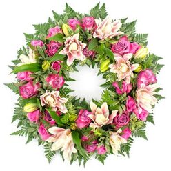 A softly radiant circular wreath, designed as a funeral or memorial tribute, is shown against a spotless white background that throws every colour and contour into sharp relief. The entire ring is packed with fresh flowers, dominated by roses in vivid tones of fuchsia, magenta, and warm pink, their petals in different stages of opening so that tight buds sit alongside full rosettes. Nestled among them are opulent double lilies in creamy white, blush, and pale peach, some fully open like starbursts, others still held in slender, green buds that promise more beauty to unfold. The blossoms rest on a bed of deep green foliage, with fine, feathery fern fronds softening the outer edge and broader, polished leaves adding structure and richness to the inner circle. Highlights of bright lime-green buds and stems create a gentle contrast to the romantic pinks, while the circular form suggests protection, eternity, and a continuous embrace. The wreath feels both lush and carefully balanced, as if arranged with quiet concentration by a local florist used to guiding North Watford families through difficult days. Under the neutral light, the textures of the petals and the delicate shadows between the leaves are clearly visible, giving the arrangement depth and presence. This is the sort of floral wreath that might rest beside a framed photograph at a crematorium chapel or accompany a hearse along St Albans Road, offering a graceful expression of sympathy and respect.