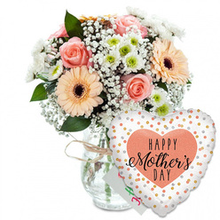 Set against a clean, uncluttered white background, this Mother's Day composition feels like a moment of quiet celebration captured in a North Watford home. In the centre stands a clear glass vase with softly curved sides, its transparency revealing a cluster of neatly trimmed stems and a faint swirl of water, suggesting freshly arranged flowers. A simple loop of natural twine is tied around the neck of the vase, giving the piece a relaxed, rustic charm that would suit a kitchen table near Garston or a terrace just off St Albans Road. Rising from the vase is a generously full bouquet in gentle pastel hues. Peach-pink roses, fully open yet still crisp at the petal edges, form the romantic heart of the arrangement. They are joined by bright peach gerbera daisies, their broad petals radiating out from deep brown centres that lend a bold, graphic contrast. Between these larger blooms, airy sprays of white gypsophila create a soft mist, while small daisy-like white chrysanthemums, each with a light green centre, add texture and volume and echo the freshness of spring walks around North Watford's green spaces. Threads of glossy green foliage weave through the flowers, framing them and giving the bouquet a natural rhythm. Tucked just behind the stems, only partly visible, is a small white greeting card with faint pink and green lettering that hints at a personal "Happy Mother's Day" message. In the foreground, slightly to the right, a large heart-shaped foil balloon leans against the vase. Its bright white surface is sprinkled with metallic gold and silver dots like scattered confetti, and in the middle a peach-coloured heart holds the phrase "HAPPY Mother's DAY" in contrasting black script, making the whole scene feel like a joyful, heartfelt tribute ready to be delivered to mums across North Watford.