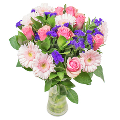 A carefully curated bouquet of blush roses and pale pink Gerbera daisies, accented with vivid violet statice, presented in a slim, transparent glass vase with visible stems and water. The pink roses range from tightly furled buds to softly opening blooms, each petal layered into a satin spiral that reflects a florist's attention to form and timing; the Gerbera daisies add cheerful roundness with light centres that seem almost luminous against the softer pinks. Clusters of purple statice thread through the arrangement, their textured, papery sprays offering a lively rhythm and a hint of wild meadow charm. Lush deep-green foliage provides a generous, cooling backdrop-leaves that appear dewy and substantial, giving weight and a woodland freshness to the top note of the composition. The lighting in the photograph is soft and natural, making the pastel palette glow while keeping the overall impression calm and elegant. Designed with everyday elegance in mind, this bouquet suits anniversaries, messages of support, or as a thoughtful surprise to lift someone's spirits on a dull afternoon. Locally sourced and arranged with care for North Watford homes and offices, this style is available through Flower Delivery North Watford and works beautifully by a windowsill on Bushey Mill Lane or as a centerpiece after a walk through Cassiobury Park-simple, refined, and reliably fresh.