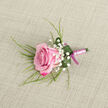 On a pale beige, linen-like fabric that offers a gentle, tactile backdrop, a meticulously crafted pink rose boutonniere becomes the clear focal point, capturing the sentiment of a wedding morning in North Watford. The rose itself is placed on a slight diagonal, its petals forming a perfect rosette that moves from a barely opened, pastel-pink centre to edges saturated with vibrant fuchsia and magenta, suggesting a bloom at the peak of its freshness. Surrounding the rose are light, cloud-like tufts of Baby's Breath, each tiny white flower adding contrast and an almost sparkling lightness, much like the soft haze of flowers you might see at a ceremony near St Albans Road. Wisps of long, slender greenery stretch outward in relaxed arcs, giving the design a sense of natural movement, while at the base, a wider, deep green leaf curls protectively around the stem cluster, its smooth surface dotted with a few dewy droplets that enhance the impression of garden-fresh foliage. The stems are bound with evident care, wrapped in a smooth satin ribbon of pale pink that hugs the stems in tight, even coils before finishing in a neat bow tied with a richer magenta ribbon. The two shades of pink on the ribbon mirror the rose's own colour transition, showing the florist's eye for harmony and detail. Set against the understated, slightly textured background, the boutonniere radiates refined romance and is perfectly suited for a groom's lapel or a formal event guest, reflecting the thoughtful floral artistry often requested for same-day celebrations across North Watford.