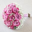 Resting gracefully on a softly textured light fabric-perhaps reminiscent of table linens in North Watford's warming homes-this round bouquet captures the essence of romantic celebration. The arrangement features an abundance of pink roses, each displaying eye-catching two-tone petals that deepen from a gentle lavender-pink at their centres to a richly saturated magenta or fuchsia around their edges. Surrounding and weaving among the roses, generous sprigs of delicate white baby's breath provide a misty, cloud-like counterpart, enhancing the airy volume and offering a gentle visual dance. The dome-shaped bouquet is masterfully bound with a soft, pearl-adorned silky pink ribbon, its shimmer recalling the romance of local North Watford weddings or anniversary soirées. Just beneath the elegant wrap, the natural stems remain visible, showcasing both the bouquet's freshness and the authenticity of its design-each stem trim and precise as only a skilled North Watford florist would ensure. Gentle diffused light brings out every textural nuance-from the velvet of rose petals to the intricate sprays of baby's breath-casting an inviting glow that speaks of new beginnings and heartfelt joy. Perfect for a bridal bouquet, an engagement, or a centrepiece for cherished moments, this arrangement embodies both grace and timeless local charm, cherished by those who prize artistry and warmth in North Watford's thriving floral scene.