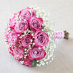 Set against a softly crumpled, light beige fabric that feels like fine linen laid out in a North Watford townhouse, this bouquet stretches diagonally across the frame with quiet confidence. At its centre, approximately ten to twelve roses glow in layered tones of lavender-pink and mauve, their inner petals cool and silvery before deepening into vibrant raspberry and magenta around the edges. Each rose is full and plush, the petals curving in gentle spirals that suggest both poise and passion. Surrounding the roses is an airy collar of white baby's breath, thousands of tiny Gypsophila blossoms creating a frothy, cloud-like border that softens the stronger pink hues and adds a touch of lightness, like early morning mist along St Albans Road. A few glossy deep green leaves emerge discreetly between blooms, giving the design a grounded, garden-inspired feel. The stems are neatly gathered and angled toward the lower right, wrapped in a smooth light purple satin ribbon that lies snug against the hands when held. Over this ribbon, a fine strand of white pearls coils delicately, providing a hint of classic bridal jewellery and ceremonial grace. The lighting is gentle and natural, enhancing the gradient of colour in each petal and the texture of the tiny white flowers. This detailed composition evokes devotion, making it ideal for wedding imagery, engagement celebrations, or romantic features focused on North Watford flower delivery and intimate local ceremonies.