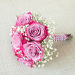 On a pale, subtly woven fabric background, a carefully crafted bouquet lies at an elegant angle, as though it has just been gently set down after photos at a North Watford registry office. The main focus is a cluster of bi-colour roses, each one large and velvety, with petals that begin in a soft lavender or light purple at the centre and gradually intensify into a vivid fuchsia and magenta pink along the edges. This natural gradient makes every bloom look painted by hand, drawing the eye into its spiralling heart. Between the roses, delicate sprays of white baby's breath form a light, cloud-like halo, their many tiny blossoms creating texture and a feeling of airiness that keeps the bouquet from feeling heavy or formal. The stems are bound firmly together, wrapped in a deep purple satin ribbon with a satin sheen that catches the light, and then embellished by a swirling strand of white faux pearls that twist down the handle like a bracelet. This pearl detail suggests care and tradition, echoing the kind of jewellery and hair accessories often chosen by brides and bridesmaids around North Watford's churches and hotels. The overall palette remains cool and romantic against the warm beige backdrop, with the roses and gypsophila standing out clearly, every petal and bead visible. The bouquet's size and style make it perfect as a bridesmaid's posy, a prom bouquet for a local school leaver, or a cherished bouquet for a small, heartfelt ceremony, capturing the quiet elegance and emotion of intimate celebrations in the area.