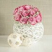 A serene indoor still life shows a lush, rounded bouquet of soft pink roses and delicate white baby's breath resting in a sculptural white vase, styled as if on a calm afternoon in a North Watford living room. Around fifteen to twenty roses are gathered tightly into a dome, each bloom unfurling from a pale blush centre into deeper pink and almost fuchsia edges, creating a gentle ombré that feels romantic but not overpowering. Between the roses, clusters of tiny white gypsophila float like clouds, softening the lines of the bouquet and adding a sense of lightness, as if the flowers have just been arranged for a birthday tea or quiet anniversary at home. The arrangement sits in a spherical vase crafted from interwoven white strands, reminiscent of painted wicker or rattan; its open, criss-cross pattern lets light filter through and throws subtle shadows on the smooth white surface beneath. To the left of the vase, two decorative spheres made from pale natural fibres echo the woven texture, one larger and one smaller, adding a minimalist, Scandinavian-style accent that would not look out of place in a modern flat off St Albans Road. The background is a softly textured wall in warm beige or cream, which keeps the focus on the gentle pinks and whites of the flowers while bathing the whole scene in calm, diffused light. The overall effect is of freshness, quiet luxury, and everyday elegance-an arrangement you might order for same-day delivery in North Watford to brighten a hallway console, coffee table, or bedside in a subtle, heartfelt way.
