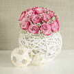 In this refined wedding floral piece, a dome of pink roses and baby's breath sits poised in an artistic white orb, as though it were designed to bring a sense of calm luxury to a North Watford reception table. Each rose is carefully nestled against the next, their petals unfolding in spirals that move from the palest blush in the heart to a more saturated pink at the outer edges, creating a soft ombré effect that looks almost hand-painted. Wisps of white gypsophila, or baby's breath, gather at the base and weave between the roses like tiny stars, giving the arrangement a weightless, cloud-like border. The flowers rise from a distinctive round vessel made of interlaced white strands, a woven globe whose open structure lets light filter through, throwing gentle, circular shadows on the surface beneath. This sculptural container, so different from a standard vase, strikes a balance between rustic handcraft and sleek modern design, a style often requested by couples marrying near Cassiobury Park or hosting intimate gatherings in North Watford's small hotels and restaurants. The setting in the image is softly lit, suggesting natural daylight from a nearby window, which enhances the tenderness of the pink tones and the purity of the white accents. The mood is serene, almost spa-like, perfect for moments when guests pause to admire the table flowers and feel a quiet sense of romance. It is easy to imagine this arrangement greeting guests at a wedding breakfast, its gentle fragrance adding an extra layer of welcome.