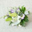 This elegant posy rests on a neutral, off-white linen surface, composed with the practiced hand of a local florist. White Oriental lilies provide the striking focal point: some blooms open fully to reveal their sweeping, curved petals, orange-yellow stamens, and tiny dark pollen flecks, while others remain as plump green buds promising more bloom. Nestled among them are lisianthus (eustoma) in white, each petal edged in lilac and violet that deepens toward the ruffled margins, creating a refined watercolor effect. Airy sprays of baby's breath (Gypsophila) thread through the bouquet like soft mist, offering delicate contrast to the glossy, broad leaves-probably ruscus or salal-and the varied greenery of feathery fronds and slim spike grasses that introduce movement and depth. The stems lie bound with a crisp white satin ribbon punctuated by pearl-like beads and subtle gemstone-style pins, a finishing touch often requested for bridal bouquets around Cassiobury Park and Watford High Street. There is a gentle interplay of textures: the smooth lilies, the frilled lisianthus, the feathery fillers, and the linen's tactile weave beneath. A faint, spicy lily scent mingles with fresh green notes, suggesting a bouquet that feels both celebratory and serene. As prepared by Flower Delivery North Watford, this arrangement reads as a classic choice for weddings or thoughtful gifts delivered across the North Watford neighbourhood.