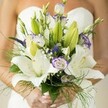 This North Watford bridal bouquet is the very essence of tender anticipation and quiet celebration, held lovingly in hands that glow with the softest hint of nail lacquer. Illuminated by gentle daylight, the bouquet reveals creamy white Oriental lilies, their petals broad and lustrous, some open in full majesty, others still tightly wrapped with promise. Pale lavender-edged lisianthus blooms swirl among the lilies, their ruffled softness suggesting windswept wildflowers glimpsed on breezy walks near North Watford parks. Deeper, almost regal, purple Veronica and dreamy clusters of baby's breath interlace amid the arrangement, imparting both depth and lightness-a bouquet alive with harmonious balance. Foliage in rich, layered greens, including firm lily buds and wispy asparagus fern, cascades around the flowers, evoking freshness and a sense of movement. The bride's white, strapless dress forms a gently blurred background at the edge of the image, keeping the viewer's attention on the rich textures and hues within the bouquet. Altogether, this arrangement is a poetic blend of tradition and natural splendour, evoking the optimism and elegance cherished by North Watford couples as they embark on a new chapter, whether celebrating in a local parish hall or beside the tranquil waters of Watford's canals.