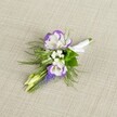 A meticulously crafted wedding boutonniere rests on a softly textured light beige linen background, the neutral cloth catching gentle, even light that makes every petal and ribbon edge easy to see. At the heart of the design sit two graceful lisianthus blooms, their petals a crisp, creamy white with deeply ruffled edges brushed in rich purple, as though they've been hand-painted. Around them, tiny white filler flowers reminiscent of baby's breath create a light, cloud-like halo, softening the contrast and adding a delicate, airy feel. A slender vertical spike of deep purple muscari, with its tight cluster of tiny bell-shaped florets, lends a sculptural line and a deeper, cooler shade of violet, echoing the lisianthus edges while bringing its own texture. Two pale green, unopened buds angle gently from the cluster, suggesting freshness and the promise of blooms still to come. Fine, feathery greenery, similar to asparagus fern, fans out behind the flowers, its soft fronds giving a sense of natural movement, as if the stems had just been gathered from a quiet corner of a North Watford garden near Cassiobury Park. The stems are gathered neatly, wrapped in smooth white satin ribbon that gleams subtly against the linen, and finished with a small, tidy bow that signals this piece is ready to be pinned to a lapel. The overall impression is one of calm refinement and gentle romance, ideal for a groom or groomsman preparing for a ceremony at a local North Watford venue, or for anyone seeking a quietly elegant floral accent for a prom or special evening. The image reveals the care and craftsmanship of an experienced florist, from the balanced colour palette to the thoughtful mix of textures and forms.