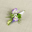 There is a restrained joy to this boutonniere, a small composition that balances purity and colour with the kind of care we bring to every piece for clients around North Watford. Two lisianthus blooms-white with soft violet edges-rise as the focal point, their layered petals cool and satin-smooth to the touch. Around them, clusters of baby's breath scatter like tiny pearls, their fine white sprays lending an almost cloud-like lightness that softens the silhouette. Below, compact spikes of grape hyacinth provide a clear blue-violet contrast, the tiny bell-shaped florets adding a lively texture that catches the eye against the dark green of feathery ferns. The stems are gathered with a white satin ribbon, its neat knot echoing the buttoned precision of a groom's lapel; the boutonniere sits on neutral linen where the woven texture deepens the sense of handcrafted simplicity. The scent is subtle-green, faintly sweet-and the whole piece feels suited to slow morning ceremonies, portraits on Cassiobury Park lawns, or milestone photographs taken near North Watford station. Prepared with the same attention to proportion and freshness that defines Flower Delivery North Watford, this boutonniere is a compact narrative of spring: tactile, fragrant, and quietly celebratory, meant to enhance a suit or jacket without overshadowing the moment.