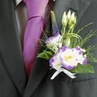 A sharply focused close-up shows the upper torso of a man dressed for a wedding or formal celebration in North Watford, the camera lingering on the rich detail of his attire rather than his face. He wears a beautifully tailored, dark charcoal grey suit jacket with a clean, structured lapel that catches the soft indoor light, and beneath it a pale grey checkered dress shirt that adds a gentle pattern without overwhelming the look. At his neck sits a luminous, magenta silk tie, knotted with care, its smooth sheen echoing the joyful occasion. Pinned firmly to the left lapel is the true focal point: a handcrafted boutonniere featuring two white lisianthus blooms edged with deep violet-purple ruffles and bright yellow centres, their petals slightly cupped as if mid-breath. Around them nestle slim, unopened white buds, tiny clouds of white filler flowers reminiscent of baby's breath, and a single upright spike of dark indigo veronica giving graceful height. Wisps of feathery greenery and broader emerald leaves frame the flowers, while fine loops of decorative green grass add a modern flourish, like stylised stems seen in Cassiobury Park flowerbeds. At the base, a small white satin ribbon is tied into a perfectly neat bow, its soft, matte texture contrasting with the crisp fabric of the jacket. The whole composition feels like a quiet promise of love and celebration, the kind of refined, carefully balanced buttonhole a local North Watford florist would create to accompany a groom walking into a church off St Albans Road or a civil ceremony at a nearby venue.