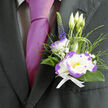 Against the charcoal weave of a smart suit, this boutonnière feels like a careful, considered gesture. Its centrepiece is a white lisianthus, the soft petals edged in a lively purple that throws a delicate, painterly shadow across the lapel. Complementary textures-slender grassy loops, dark glossy foliage, and tiny sprays of baby's breath-frame the bloom and create a sense of purposeful movement; a single upright spike of indigo veronica punctuates the composition with a vertical accent that lifts the eye. Three pale lisianthus buds sit just behind the main flower, a quiet echo of new beginnings, while subtle green buds and blades suggest freshness and vitality. A neat white ribbon holds the stems together with clean, understated elegance, and the whole piece rests beside a magenta silk tie, the contrast giving personality without overpowering the suit. In the soft light, petal edges shimmer and the purple rim adds definition and drama, a tonal detail that speaks to careful selection and skilled handling. As a local florist familiar with deliveries to the area around Watford Junction and small ceremonies off Hempstead Road, I often recommend this style for weddings, anniversaries, and commemorative events; it is intimate enough for a best man yet refined enough for a groom. Its gentle scent and tactile softness offer steady comfort through long days of celebration, leaving a subtle, lasting impression of tenderness and thoughtful craft.