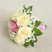 A small but exquisitely detailed bouquet is photographed from a slightly elevated angle, lying diagonally across a neutral light beige fabric that has the subtle, tactile texture of linen or fine burlap, like something seen on a carefully laid table in a North Watford townhouse. At the centre of the design sit five creamy white roses, their petals velvety and gently unfurling, each blossom full yet soft-edged, giving a sense of calm and classic romance. Just to the front-left of this cluster, a single rose in bright fuchsia pink commands attention, its saturated colour adding drama and individuality against the ivory tones. Threaded among the roses are delicate clouds of baby's breath, the tiny white flowers floating on slender stems and creating a lace-like veil that lightens the whole composition. Around the outer edge, glossy deep green leaves, likely ruscus or salal, extend in pointed shapes, grounding the bouquet with a fresh, natural contrast. The stems are neatly tied and wrapped in a bold pink satin ribbon, smooth and slightly reflective, forming an elegant handle that suggests it could be worn or carried. Over the ribbon, slim rows of small, iridescent white pearls run horizontally, catching the light like little beads of dew and giving the piece a refined, ceremonial touch. The lighting is soft and even, with no harsh shadows, enhancing the gentle mood. Overall, the bouquet feels perfectly sized for a corsage, boutonnière, or small centerpiece for a North Watford celebration-perhaps a wedding near St Albans Road or prom photos by the local green-carrying an air of romance, attention to detail, and quiet joy.