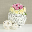 This image presents an elegant, close-up still life of a meticulously crafted rose arrangement, the kind a local North Watford florist might prepare for a special family gathering near St Albans Road. Filling the centre of the frame is a neat, spherical bouquet composed of lush pink and creamy pale yellow roses, each flower carefully placed so the colours intermingle in a soft, harmonious rhythm. The petals are plump and velvety, some tightly cupped, others just beginning to unfurl, suggesting freshness and the promise of fragrance. Interwoven among the roses are wisps of delicate white baby's breath, forming a floating veil of tiny blossoms that soften the edges of the bouquet and lend it a romantic, almost bridal quality. A hint of green foliage is visible at the base of the flowers, tucked in sparingly so the roses remain the star. Supporting the bouquet is a distinctive white orb-shaped vase, designed from slender, crisscrossing rattan-like strips that create an airy, open structure, reminiscent of a hand-woven nest. This sculptural vessel rests on a bright white surface, flanked on one side by two decorative spheres formed from natural beige wooden strips, echoing the same woven texture and adding a subtle, rustic counterpoint to the polished arrangement. Behind it all, a soft beige background with a faint linen effect stretches across the frame, its neutral tone allowing the pastel colours to glow gently. The overall mood is serene and contemporary, ideal for a wedding reception table, an understated anniversary gift, or an elegant centrepiece for a freshly redecorated North Watford home, with the quiet suggestion that a local delivery could bring this scene directly to a loved one's table.