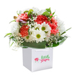 There's a gentle yet exuberant spirit to this bouquet, expertly arranged and displayed in a pristine white gift bag-a signature touch often found in North Watford's favorite local florists near Langley Road. Coral and soft peach carnations, their ruffled petals brimming with both vibrancy and subtle fragrance, mingle closely with cheerful white daisies, their lime-yellow centers glowing like warm spring sunshine. Clouds of delicate baby's breath add an ethereal softness, surrounding the main flowers in a light, airy embrace, while shy buds tinged with purple nestle quietly on the arrangement's left, hinting at nature's unfolding surprises. Deep, polished green leaves envelope the flowers, forming a wide, graceful collar that gives the entire bouquet strength and compositional balance-much like the green borders that define North Watford's parks. The bouquet is shaped into a generous, rounded mass, each bloom carefully angled to foster a sense of abundance and thoughtful intention. Bright, diffuse lighting accentuates the freshness and true-to-life colour palette, isolated elegantly against the white backdrop. Finished with sturdy white ribbon handles, the sleek bag underlines the bouquet's readiness for gifting, perfect for celebrating birthdays, milestones, or simply bringing a heartfelt smile to someone in North Watford. Every detail, from the flower choices to the careful presentation, reflects a florist's artistry and a community's warm spirit.