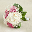 Set against a calm, light beige fabric that looks like finely woven linen, this hand-crafted bouquet unfolds horizontally like a still-life painting created for a North Watford wedding album. In the centre blooms a generous creamy white peony, its many layers of ruffled petals opening softly, as if caught at the perfect moment between bud and full flourish. Around it, rich fuchsia-pink roses and softer blush-toned roses cluster closely, their spiralled hearts and velvety texture bringing a sense of passion and devotion to the composition. Wisps of white gypsophila weave gently through these main blooms, the tiny star-like flowers forming a delicate haze that lightens the bouquet and adds an almost sparkling texture, like morning mist over Callowland and the nearby gardens. Deep, glossy green leaves curl gracefully around the outer edge and base, cradling the flowers while giving the arrangement a confident, sculpted outline. The stems are neatly aligned and wrapped in a clean white satin ribbon, creating a smooth, elegant handle ready to be held by a bride walking through a North Watford church doorway or stepping into a registry office celebration. Soft, diffused lighting washes over the bouquet, highlighting the interplay between matte petals and shiny foliage, and casting the faintest shadows that add depth without drama. The overall effect is tranquil yet emotionally charged, evoking early spring or summer mornings and the gentle promise of a shared future. It feels like a bouquet designed by a local florist who understands the quiet, heartfelt moments behind every celebration in North Watford.