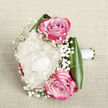 On a pale, linen-like backdrop in soft beige tones, a compact hand-tied bouquet is captured from a slightly raised angle, revealing a careful balance of colour, texture, and form. The heart of the arrangement is a large creamy white peony, its petals layered in generous ruffles, creating a pillowy, almost cloud-like bloom that exudes quiet sophistication. Flanking this central flower sit two rich pink roses, one poised a touch higher and one lower, each rose swirling inward to a tight centre before opening outward into petals that shift from gentle blush to a deeper fuchsia edge. Threads of baby's breath, with their clusters of tiny white flowers, wind delicately between the larger blooms, adding an ethereal, misty quality that softens every line. Around the base, dark green aspidistra leaves are folded and wrapped like a protective collar, their smooth, glossy surfaces contrasting with the softness of the petals. The stems are firmly gathered and finished with a smooth white satin ribbon, emphasising that this is a bouquet meant to be held for a ceremony, perhaps carried down the aisle at a North Watford wedding near St. Albans Road or delivered to a loved one in the nearby residential streets. Gentle, diffused light glows across the arrangement, highlighting each petal edge and the subtle shadows within the peony's centre. The overall impression is one of timeless romance and graceful celebration, ideal for couples seeking classic wedding flowers or a meaningful, hand-delivered gift in and around the North Watford area.