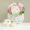 At the centre of the scene, a lush, dome-shaped bouquet draws the eye, its creamy white peonies and bold fuchsia-pink roses nestled tightly together like a cluster of soft clouds and jewels. The peonies are full and generous, their many petals unfolding in velvety layers, while the roses offer a stronger, more defined shape, their vibrant pink tones suggesting passion and joy. Fine strands of baby's breath thread through the composition, their tiny white blossoms creating a delicate mist that softens the transitions between each larger flower. Around the base, glossy green leaves fan out and curve gently, grounding the bouquet in natural freshness and echoing the greenery found in the parks and gardens around North Watford. The arrangement sits in a striking white vase crafted from slender, intertwined strands that form an open cage, giving the impression of a modern wicker sculpture rather than a traditional container. Through its lattice, you can glimpse shadows and hints of stems, adding visual intrigue and a sense of lightness. On the smooth white surface in front of the vase, two decorative orbs made from pale, woven material rest side by side, their open structure mirroring the vase's design and creating a cohesive, contemporary tablescape. Behind it all, a neutral beige background with subtle texture provides a calm, unobtrusive setting, much like a softly lit wall in a North Watford town house or wedding venue. The lighting is bright yet gentle, highlighting each petal and weave without harshness, making the arrangement feel ideally suited to a wedding breakfast table, an engagement celebration, or an elegant anniversary dinner locally.
