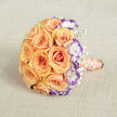 This bouquet lies as if set down after a whispered exchange of vows, its rounded form resting on a warm, beige linen that reads like a small stage for the blooms. Peach and soft orange roses form the design's heart-each petal unfurling in layered spirals that gather light and scent, evoking a gentle dawn. Framed among these, white lisianthus with vivid violet rims introduce an unexpected, elegant punctuation: their frilled edges and cool tones bring depth and a slightly contemporary edge to the classic rose palette. Wisps of baby's breath float between larger flowers, lending a sense of breath and innocence as they peek out like tiny, white constellations. The stems are wrapped with a peach-corally satin ribbon, smooth to the touch and finished with a fine string of creamy pearls that coils down the handle like a secret kept close to the heart. The textured fabric beneath picks up soft shadows and highlights, accentuating the petals' velvety surfaces and the pearls' soft glow, while the overall light is calm and natural, like late-morning sun filtered through a curtain. This bouquet carries an emotional clarity-romantic, joyous, quietly luxurious-and is ideal for intimate weddings or anniversary ceremonies. Locally, it suits bridal moments captured against the mellow greens of North Watford's parks or the gentle brickwork near Vicarage Road, making it both visually memorable and deeply appropriate for the neighbourhood's understated celebrations.