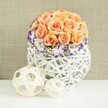 An elegant floral centerpiece sits poised on a bright white surface, evoking the kind of refined table styling often requested for weddings and special gatherings around North Watford. Centered in the composition is a perfectly shaped dome of soft peach and apricot roses, massed so closely that the eye perceives an almost seamless mound of petals. Each rose appears fully open, with gently cupped outer petals and tighter whorls in the centre, giving the impression of depth and softness. At the base of this rose globe, a ring of pristine white eustoma blossoms unfolds, their ruffled petals delicately edged with a vivid violet or purple line that adds a sharp yet graceful contrast to the warm pastel tones above. Tiny, cloud-like tufts of white baby's breath weave between the lisianthus, lending a light, airy texture that keeps the bouquet feeling fresh and romantic. The flowers are arranged in a distinctively round white basket-style vase made from woven wicker or rattan, its open lattice structure creating a contemporary, sculptural feel. To one side, two natural-toned decorative balls made from fine wooden strips lie on the same white surface, one slightly larger than the other, echoing the circular forms and woven textures of the vase. Behind, a softly textured beige fabric background, reminiscent of linen or burlap, provides a warm, neutral stage that enhances the arrangement's colours without competing with them. The overall effect is one of gentle luxury and calm celebration, ideal for dressing a wedding table in North Watford, adding grace to a small reception at a local restaurant, or creating a romantic focal point in a home dining room.