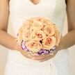 A close-up, chest-level photograph shows a bride standing quietly, her torso framed against a soft, light background that could be a sunlit room in a North Watford townhouse or a bright corner of a local registry office. She wears a traditional white wedding gown with an ivory tint, the bodice adorned with delicate lace and a gentle V-shaped neckline that leads the eye down to her waist. Her skirt falls in smooth, graceful folds, the fabric catching the light in subtle ripples that echo the calm of a late afternoon along St Albans Road. Cradled in both hands, with neatly manicured nails finished in pale polish and a hint of glitter on one thumb, is a round, meticulously crafted bridal bouquet. At its heart are numerous tightly clustered roses in soft peach, light apricot, and blush pink, each bloom densely petalled and slightly cupped, creating a lush, domed shape. The colours feel warm and romantic, like a quiet summer sunset over Cassiobury Park. Just beneath this generous crown of roses, a vivid ring of deep purple flowers with ruffled white edges, likely lisianthus, creates a dramatic collar that gently peeks out, adding contrast and depth without overpowering the centre. Tiny sprigs of white filler, reminiscent of baby's breath, lace through the base, giving the arrangement an airy, almost cloud-like softness. There is no visible foliage beyond the minimal greenery that supports the blooms, allowing the pastel roses and violet accents to remain the undisputed focus. The overall impression is one of poised elegance and tender anticipation-a bouquet designed for a bride about to walk down the aisle in North Watford, captured at the quiet, breath-held moment before the day truly begins.