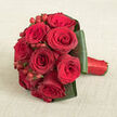 A luxurious bouquet of deep red roses is arranged in a classic rounded cluster, lying gracefully on a warm, light beige fabric that looks like softly textured linen. The roses are the star of the design, each head tightly packed beside the next, their velvety petals spiralling inward from dark crimson outer edges to rich, almost wine-red centres. Tiny, spherical hypericum berries, shining in a brighter cherry red, are scattered thoughtfully amongst the blooms, creating moments of contrast and a playful rhythm within the otherwise smooth sea of petals. Encircling the roses is a precise collar of broad, dark green leaves, meticulously folded and layered so they sit flat and secure, lending both structure and a contemporary, tailored outline. The handle of the bouquet stretches to the right, bound in vivid red satin ribbon that catches the light and reinforces the sense of cohesion and care in the design. The soft, neutral backdrop allows the reds and greens to command full attention, and the gentle overhead lighting picks up every curve, edge, and glint of moisture on the petals. This is the sort of romantic, statement bouquet you might carry at a registry office wedding near North Watford station, or present as a deeply heartfelt gift for a milestone anniversary celebrated at home. It feels intimate yet grand, like something created early in the morning in a local North Watford workshop, destined to bring a flush of emotion the moment it is placed into someone's hands.