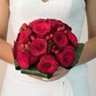 Brought together by the skilled hands of a North Watford florist, this close-up photograph reveals a lush, rounded bridal bouquet celebrated for its colour and artistry. High-quality deep red roses form the bouquet's heart, each bloom in a unique stage of unfurling-some buds promising secrets yet to blossom while others display broad, velvet petals in luminous, passionate shades. Between the roses, clusters of ruby-red hypericum berries emerge, gleaming like small gems and bringing playfulness and depth to the bouquet's composition. The entire creation is encircled by broad, deep green foliage, smoothly wrapped to provide structure and a harmonious finish, all gently held by a bride wearing a classic, lace-detailed white gown. The soft background glow accentuates the bouquet's vividness, making the reds more intense against the creamy tones of the bridal attire-a vision familiar to couples choosing local, bespoke flowers for ceremonies near North Watford's iconic green spaces. This bouquet speaks of tradition, devotion, and joy, a fragrance wafting in the imagination-fresh, romantic, and unmistakably crafted for a North Watford celebration.