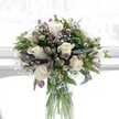 A softly lit floral arrangement stands in a clear, cylindrical glass vase, positioned as if on a bright windowsill in a calm North Watford home. At the heart of the bouquet, creamy white roses are fully open, their velvety petals spiralling outward in perfect, romantic rosettes that catch the gentle daylight. Threaded between them are clusters of lavender and muted purple alstroemeria, some buds still slender and closed, others just beginning to unfurl, their petals streaked with deeper violet veining that adds quiet drama. Tiny stars of white baby's breath drift like a delicate mist between the larger blooms, creating a cloud-like softness that lightens the entire design. Around the edges, lush green foliage in varying shapes and tones-from broad, polished leaves to more slender, tapering stems-frames the flowers and gives the bouquet a rounded, balanced silhouette. Through the transparent vase, the flower stems are neatly aligned and immersed in clear water, showcasing the natural structure and fresh, just-cut quality of the design. The whole scene feels serene and hopeful, like a still moment on a spring morning walk through Cassiobury Park before the day begins. This bouquet looks ideal for placing on a dining table in a terraced house near St Albans Road or on a bedside cabinet in a quiet flat off Garston Lane, bringing a mood of purity, comfort, and gentle celebration. Its graceful palette of whites and soft purples suggests thoughtful sympathy, a peaceful get-well wish, or a tender congratulations, each bloom arranged with the care of a florist who understands how flowers can speak when words are hard to find.
