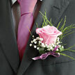 Pinned to the lapel of a dark, tailored suit, this blush-pink rose boutonniere radiates a quiet, timeless romance that would suit any North Watford wedding or prom. The rose is fully opened yet still fresh, its velvety petals layered in a soft spiral of pale shell pink that deepens just slightly toward the centre, as if the colour has pooled there. Surrounding it, airy stems of white gypsophila drift like tiny clouds, giving the design a dreamy lightness and echoing the gentle excitement of a special day. Slender, arching greenery, perhaps a refined fern or aspidistra loop, curves around the bloom, adding just enough structure to make the rose stand out against the charcoal fabric of the jacket. At the base, a slim ribbon in a muted lilac or mauve tone has been carefully wrapped and tied, the satin finish glimmering where the light touches it, much like evening sun on the brickwork of North Watford terraces. The workmanship is precise but never stiff: stems are trimmed cleanly, the proportions balanced so the boutonniere nestles close to the heart without overwhelming the lapel. It feels like the kind of floral accent chosen after thoughtful conversations with a local florist who understands how important these small details are, whether the wearer is saying vows, arriving at a family celebration, or posing for photographs near the local town hall. Seen up close, every element whispers of tenderness, commitment, and the joyful nerves of a moment that will be remembered for years.