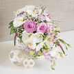 A graceful basket arrangement of wedding flowers is displayed on a smooth, reflective white table, the scene composed with the calm precision of an experienced North Watford florist. The white, cross-hatched wicker basket overflows with soft romance: full, blush-pink roses unfurl in lush, velvety layers, mingling with arching stems of white Phalaenopsis orchids whose wide, satiny petals and lemon-touched centres lend a refined, contemporary edge. Around these focal blooms, white lisianthus blossoms-some tinged delicately with lilac-open among sprays of airy gypsophila, like wisps of cloud captured in a bouquet. Smaller, dusky-pink flowers, reminiscent of stock or Sweet William, help create an asymmetrical cascade, draping gracefully over the basket's edge. A narrow spire of deep purple-magenta Veronica stands slightly taller, adding a slender, linear accent that contrasts beautifully with the rounded roses and orchids. Lush green foliage snakes through the design, from streamlined leaves to tiny buds, bringing depth, texture, and the feeling of fresh garden growth you might see on a morning walk near Cassiobury Park. At the base of the arrangement, two light, wooden-look rattan spheres sit on the glossy tabletop, their open, woven structure mirroring the basket's pattern and adding a simple decorative detail. The entire display is set against a softly crinkled beige backdrop, gently lit so that each petal catches the light without glare, making the whites glow and the pinks appear warm and inviting. The overall mood is romantic yet modern, ideal for bridal tables, engagement parties, or elegant home delivery throughout North Watford.