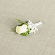 At the heart of this composition is a flawless ivory rosebud, its creamy petals arranged in soft concentric layers that invite touch and quiet admiration. Nestled close to it are tiny clusters of white baby's breath, lending a cloud-like lightness that balances the deeper tones of the greenery. Slender, feathery sprigs of dark green foliage-evocative of evergreen or cedar-reach outward, their fine texture contrasting with the broader, glossy leaves that cradle the bloom and provide a reassuring architectural frame. The stems are bound with a thin band of white satin ribbon, finished in a modest bow that suggests thoughtful restraint rather than flourish.

The boutonniere lies on a textured light-beige woven fabric that feels warm and natural in the light, a backdrop chosen to let the rose's soft cream and the foliage's rich greens read clearly in photographs. There is a restrained, almost intimate perfume: the rose's clean floral note, a faint herbal whisper from the greenery, and the barely-there sweetness of gypsophila. The overall mood is calm and sincere-an accessory designed to sit close to the heart and mark a meaningful moment.

As a North Watford florist, I imagine this pinned at small ceremonies near Cassiobury Park, during family portraits on Watford High Street, or carried into intimate gatherings by fathers and groomsmen. Prepared with care by Flower Delivery North Watford, it is a compact emblem of purity and new beginnings, suited to weddings, proms, and formal gatherings where subtlety matters.