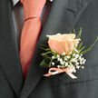 This boutonniere presents a careful study in soft contrasts: a single, impeccable peach rose sits at its heart, petals unfolding like dawn and carrying a delicate, sweet scent that seems to promise joy. Surrounding the bloom, tiny sprays of white baby's breath form an almost halo-like cushion, their cloud-like clusters introducing a light, whimsical texture. Slender green foliage and a curled support leaf give the arrangement shape and motion, their fresh, dewy tones balancing the rose's warm coral-pink glow. A pale peach satin ribbon is tied at the base in a modest bow, the sheen of the ribbon mirroring the tie's warm coral-pink and bringing a tailored finish to the piece. Against the charcoal suit lapel the colours read vivid and refined; the dark cloth deepens the rose's warmth and allows every delicate botanical detail to stand out. There's a sensorial hush to the image - the soft petal velvet, the whisper of gypsophila, the faint fragrance that floats up when the boutonniere is lifted - all speaking of attentive craftsmanship. Crafted with local care by Flower Delivery North Watford, this boutonniere suits those marking weddings or milestone celebrations across North Watford, whether walking from Clarendon Road venues or stepping out after a photoshoot in Cassiobury Park. It's an elegant, heartfelt accent that feels both timeless and intimately personal.